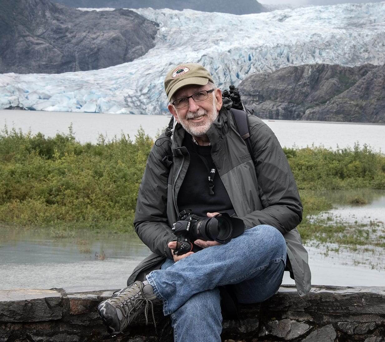 Man sitting outdoors near a glacier, holding a camera and smiling.