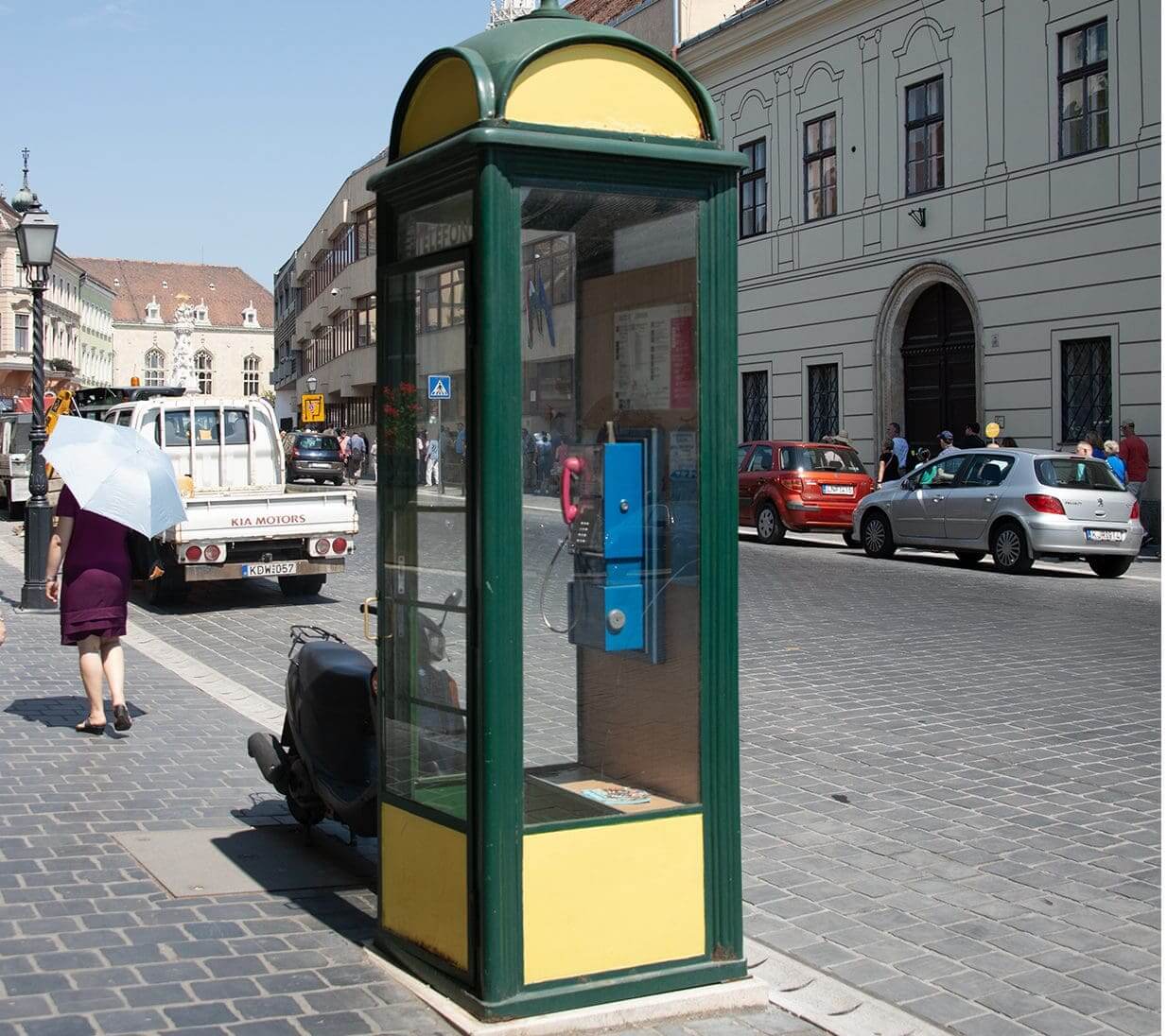 A vintage glass phone booth on a city sidewalk with cars and buildings in the background.